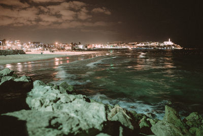 Illuminated buildings by sea against sky at night