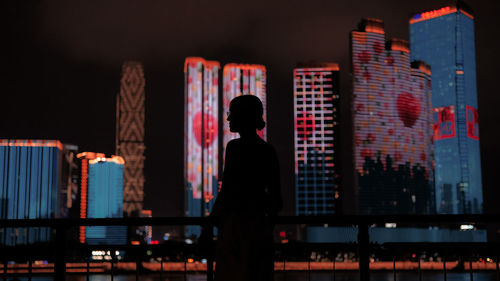 Rear view of silhouette man standing by railing against buildings at night