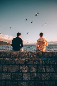 Rear view of men sitting on wall by sea against sky