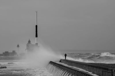 Man walking on dam while waves splashing in sea against clear sky