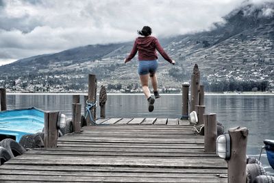 Rear view of woman jumping on pier at lake against sky