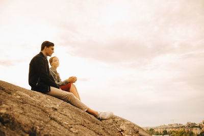 Low angle view of friends talking while sitting on rock formation against cloudy sky
