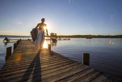 Man with paddleboard walking on pier against sky during sunset