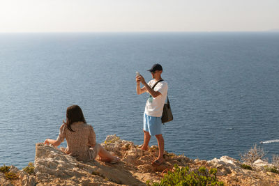 A young guy takes a photo of a girl on his phone by the sea.