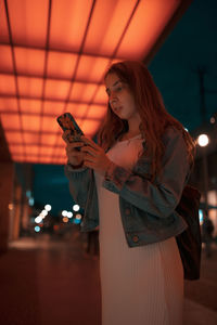 Woman using mobile phone while standing on footpath under illuminated lights