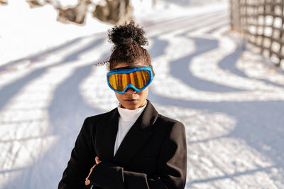 Stylish young ethnic female athlete in protective glasses looking at camera on snowy roadway in sunlight