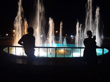 Rear view of silhouette man standing against illuminated fountain at night