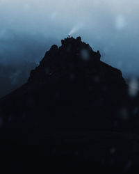 Low angle view of silhouette mountain against cloudy sky with man on top of it pointing headlight in