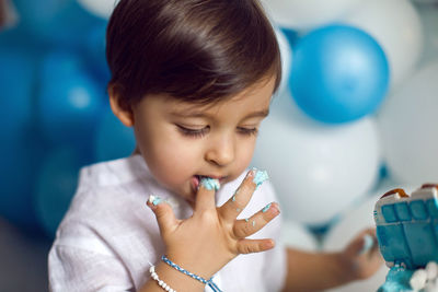 Baby boy in blue pants and shirt standing on the floor next to the cake in blue and white balloons