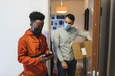 Mid adult man with package standing by male delivery person using smart phone during pandemic