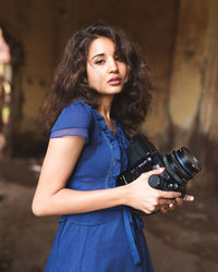 Portrait of woman holding camera while standing against wall