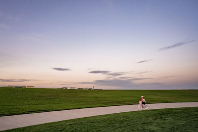 Young girl bikes down a path in a park with mowed grass at sunset