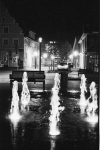 Water fountain on street against illuminated buildings in city at night during winter