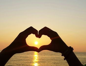 Close-up of hand making heart shape at beach against sky during sunset