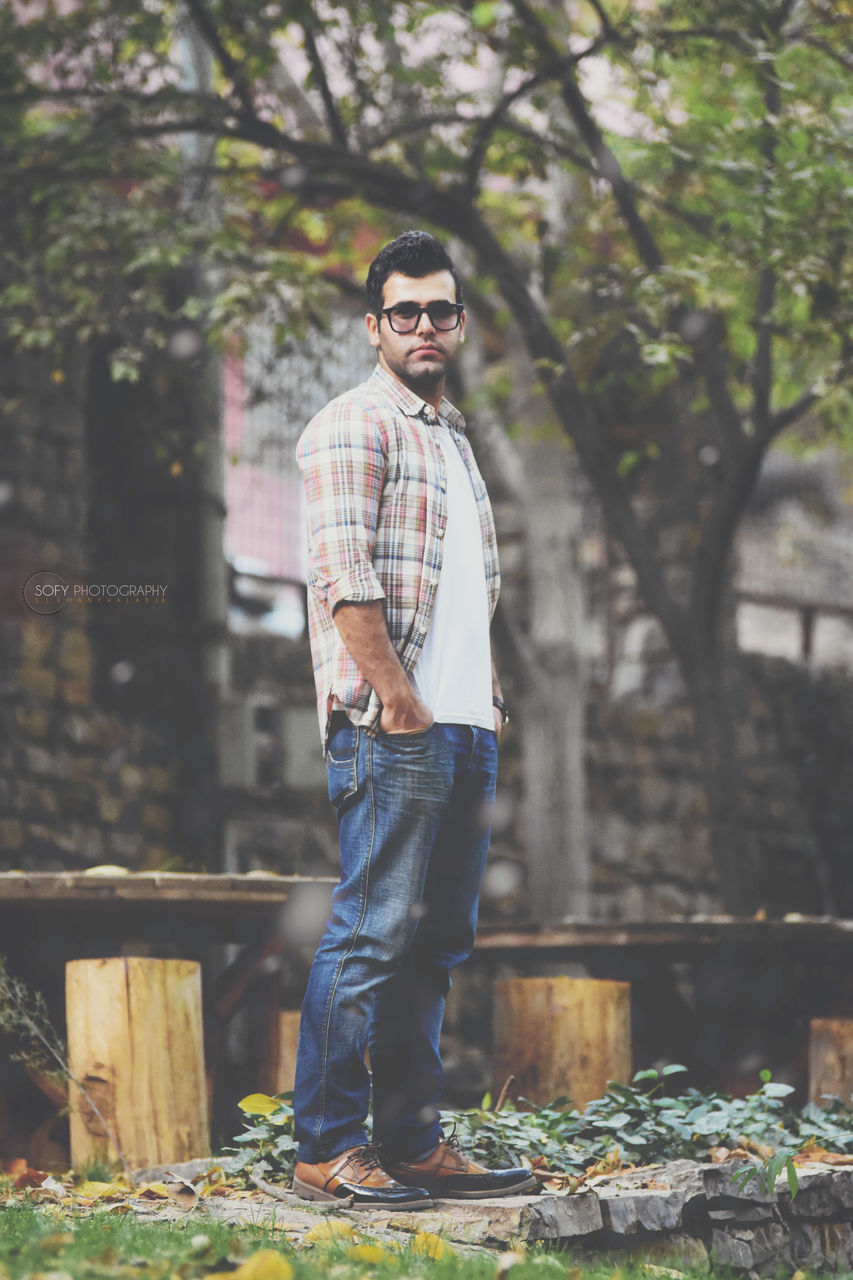standing, glasses, real people, casual clothing, one person, young men, young adult, fashion, lifestyles, tree, leisure activity, focus on foreground, sunglasses, portrait, day, plant, full length, looking at camera, nature, outdoors, jeans, plaid shirt