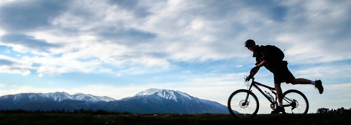 Full length of man riding mountain bike by snowcapped mountains against sky