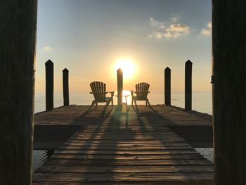 Empty chairs on pier by sea against sky during sunset