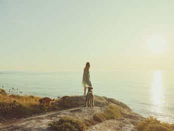 Woman standing with dogs by sea on rock during sunset