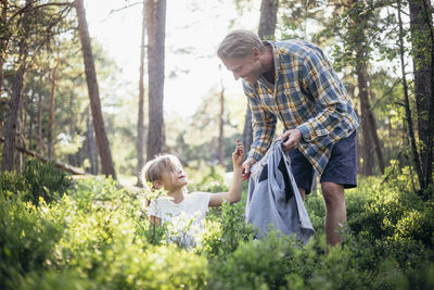 Smiling father looking at daughter collecting garbage in forest