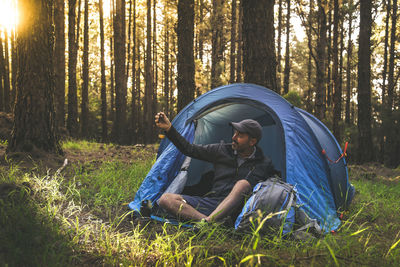 Man taking selfie while sitting in tent on field against trees at forest