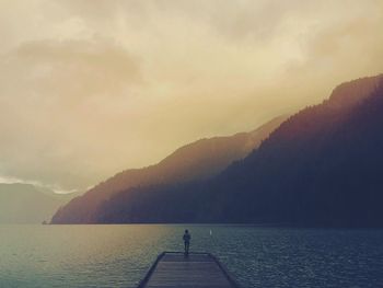 Man standing on jetty while looking at lake and mountains during sunset