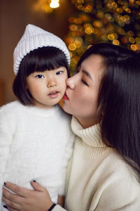 Beautiful korean mom kiss baby daughter sitting in white knitted sweaters at the christmas tree