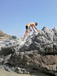 Full length of shirtless man on rock at beach against sky