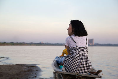 Young woman sitting in boat on shore against sky during sunset