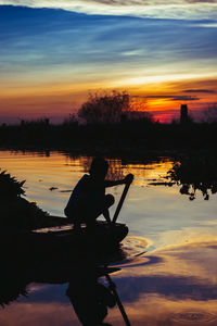 Silhouette man sitting by lake against sky during sunset