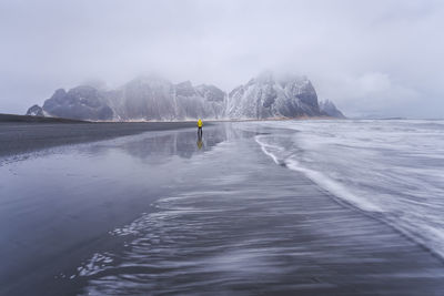 Distant view of unrecognizable traveler standing on wet black sand beach near waving sea against mountain ridge and cloudy sky in iceland