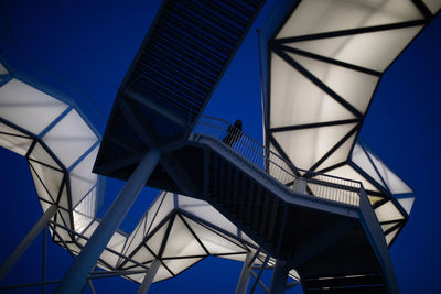 Low angle view of woman standing on footbridge against illuminated structure at dusk