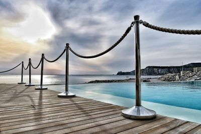 Railing on pier at poolside against cloudy sky