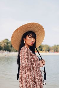 Portrait of young woman wearing hat standing at beach against sky