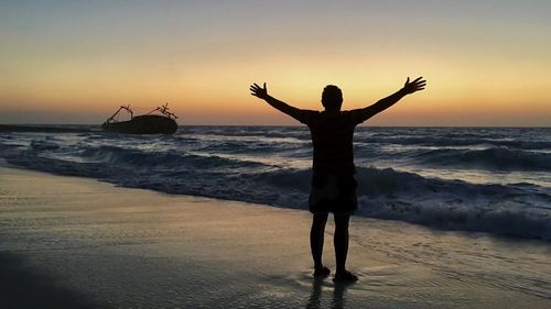 Rear view of man standing at beach against sky during sunset