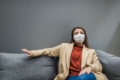 Portrait of woman wearing mask while sitting on sofa against wall