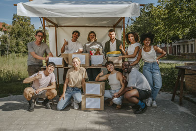 Portrait of diverse male and female volunteers showing thumbs up gesture while posing with boxes during charity drive at