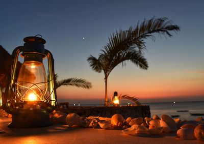 Illuminated lights on table by sea against sky at sunset