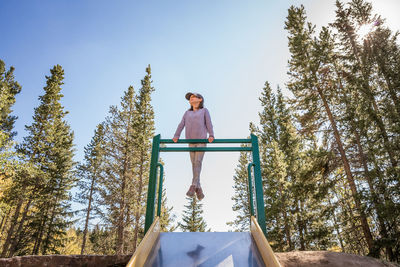 A smiling young girl plays on a playground slide among tall pine trees