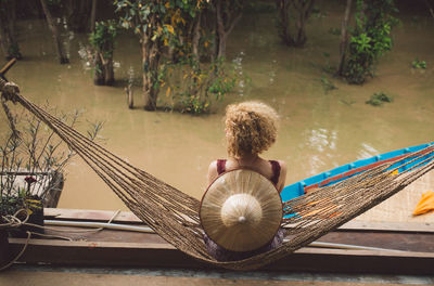 Rear view of mid adult man with hat sitting on hammock over lake