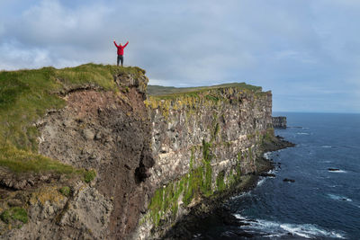 Rear view of man standing on cliff by sea against cloudy sky