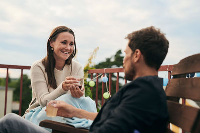 Cheerful woman talking with male friend while holding drink on terrace during social gathering