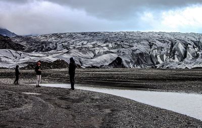 Friends standing on sand at glaciers against cloudy sky