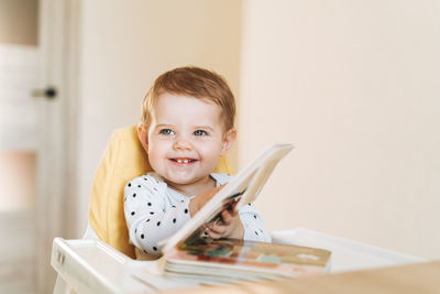 Happy cute baby girl in booster seat reading children book at home