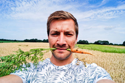 Portrait of man holding carrot in mouth on agricultural field against sky