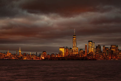 Illuminated buildings by sea against cloudy sky