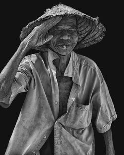 Portrait of senior man with cigarette saluting while standing against black background