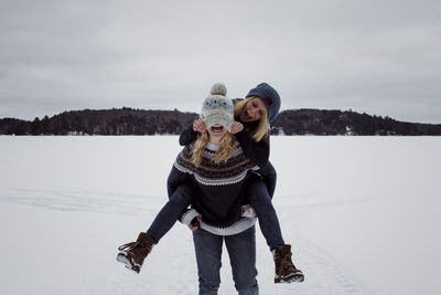 Playful woman pulling friend's knit hat while being piggybacked by her on snow during winter