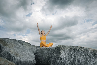 Sporty woman sitting crossed legged on rocks meditating and practicing yoga, dramatic sky.