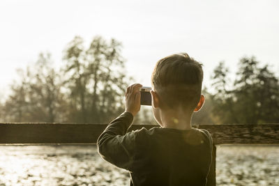 Rear view of boy photographing with smart phone against clear sky