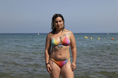 Young woman in bikini standing at beach against sky